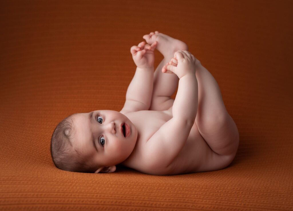 Bebé tumbado boca arriba cogiéndose los pies sobre fondo teja