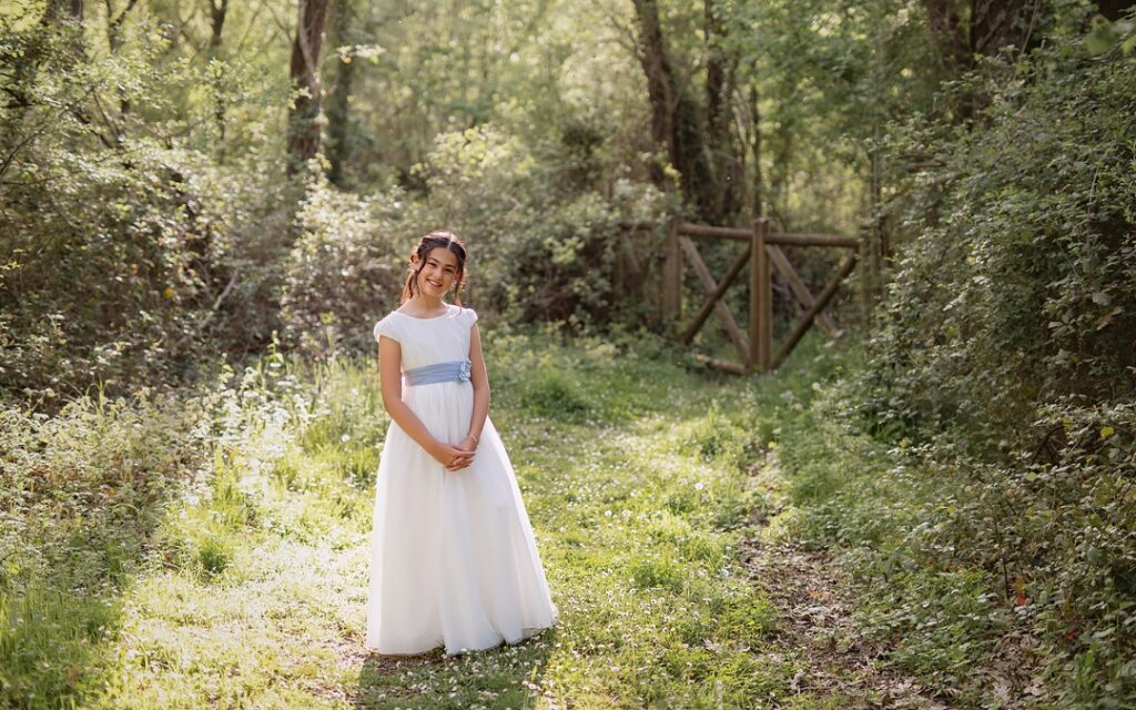 Niña con vestido de comunión blanco y fajín azul sonriendo en un claro del bosque durante su sesión de fotos de Primera Comunión