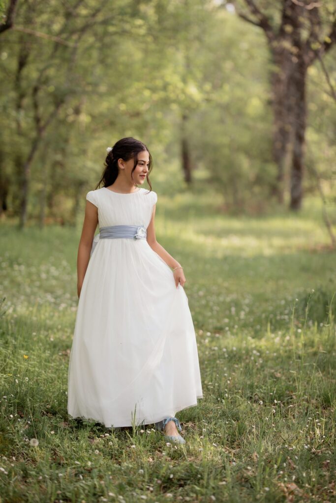 Niña paseando con su vestido de comunión por una pradera con flores en el bosque