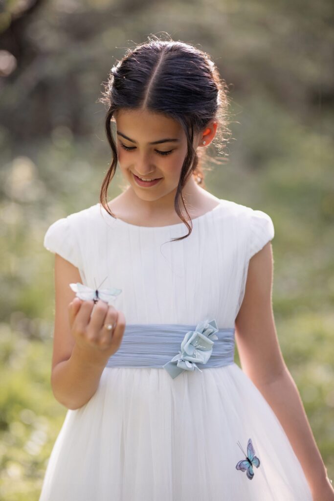 Niña mirando una mariposa en su mano con su vestido de comunión en una sesión de fotos en exterior