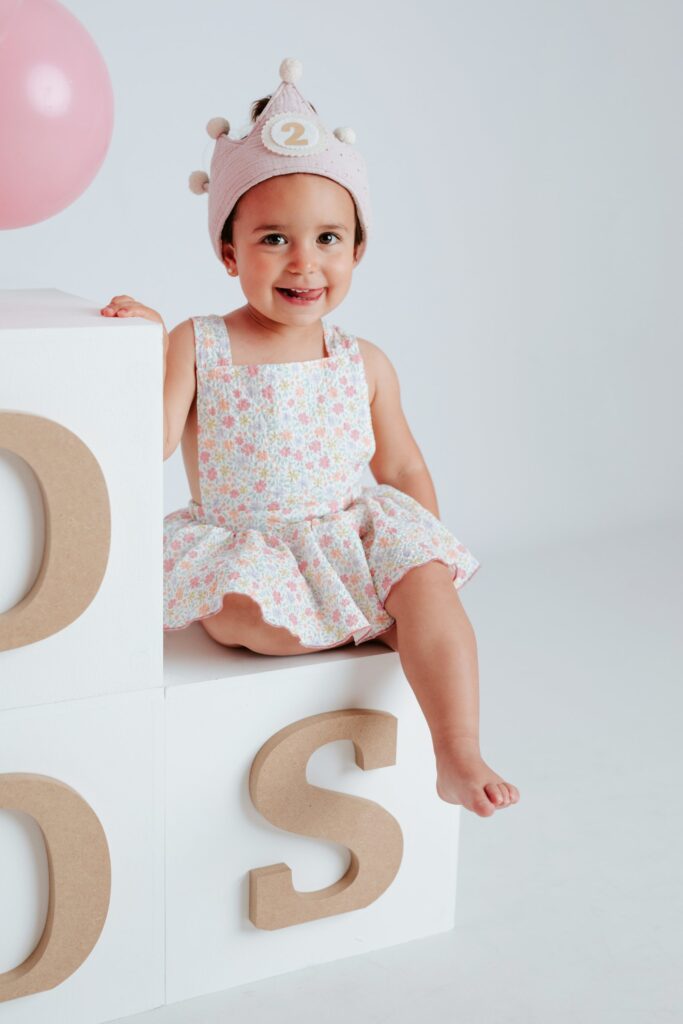 Niña sonriendo con corona y vestido de flores en su sesión de fotos de segundo cumpleaños en el estudio