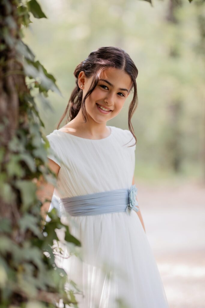 Niña sonriendo con su vestido de comunión asomada junto a un árbol con hiedra en una sesión de fotos en el bosque