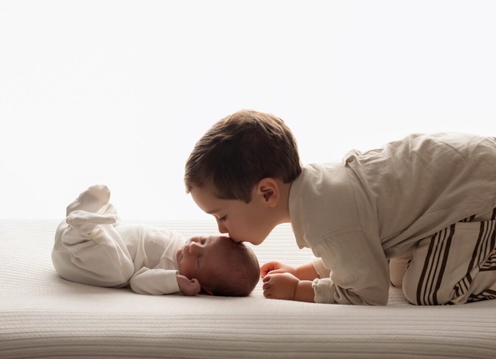 Hermano mayor dando un beso en la cabeza a su hermanito recién nacido, ambos vestidos de blanco y crema