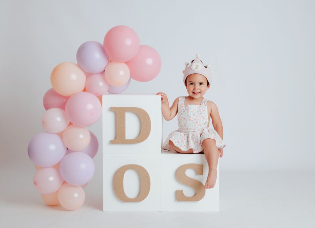 Niña de dos años con corona sentada en cubos con las letras DOS y globos rosas en una mini sesión de cumpleaños