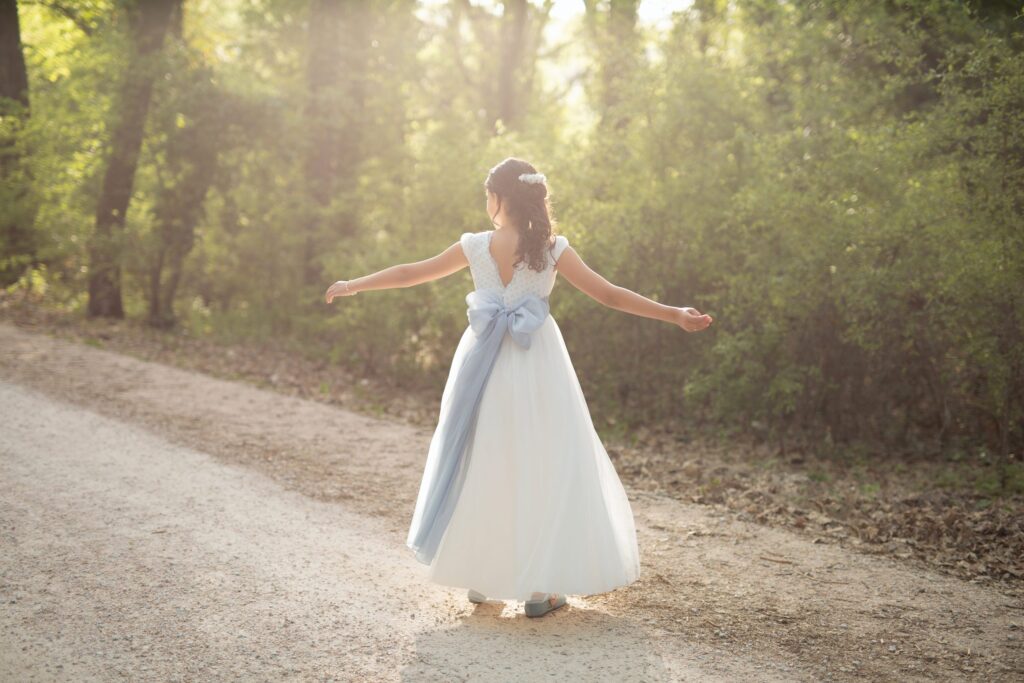 Niña de espaldas con los brazos abiertos y un gran lazo azul en su vestido de comunión en un camino del bosque a contraluz